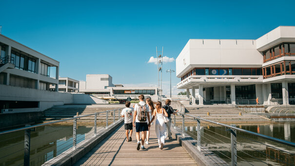 Studierende gehen auf der Brücke über den Uni-See in Richtung Zentrales Hörsaalgebäude der Universität Regensburg.
