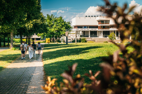 Eine Gruppe Studierender geht über den Campus der Universität Regensburg. Im Hintergrund ist die Zentralbibliothek zu sehen.