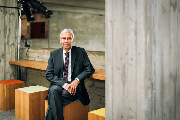 UR-Präsident Prof. Dr. Hebel sitzt auf einem der farbenfroh bezogenen Holz-Hocker auf der Galerie des Auditorium maximum