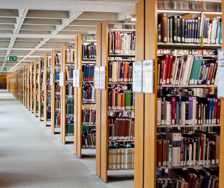 Ein langer, heller Gang, quer dazu sind bis zum Horizont mit Büchern gefüllte Holzregale zu sehen.