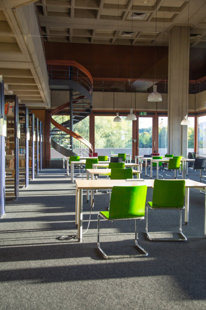 Blick in den Allgemeinen Lesesaal: Grüne Stühle vor weißen Tischen, am linken Rand sind Bücherregale zu erkennen, im Hintergrund eine Wendeltreppe und große Fensterfronten, die Ausblick auf Bäume und Rasenflächen geben. (öffnet Vergrößerung des Bildes)